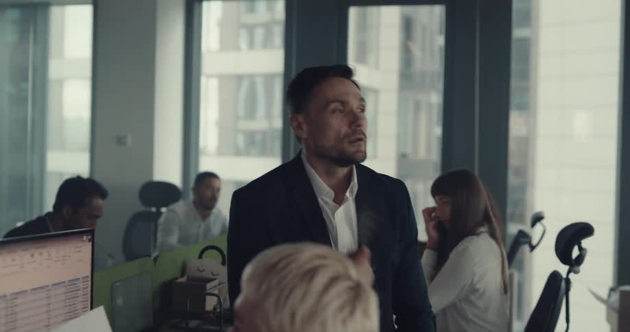 A man sits on a desk and fans himself while people work around him