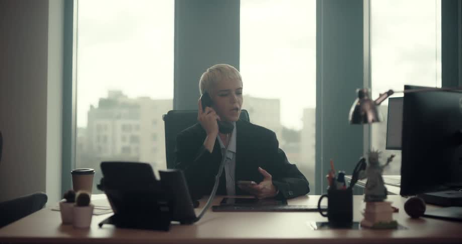 Woman sitting at desk in office answers phone and has conversation while checking her watch