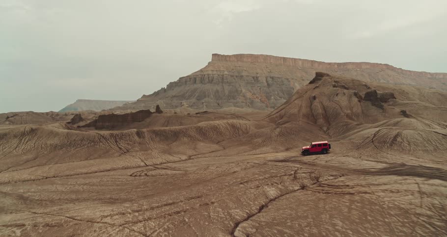 A red truck is traveling across the desert badlands with rock formations and mesa in the distance