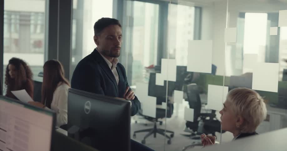 Man sits on table talking to employee regarding data on monitor in office