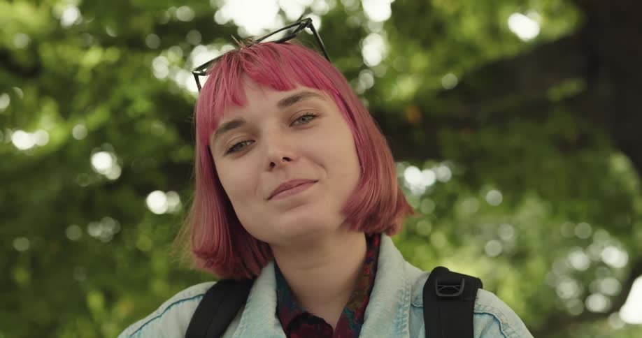 A teenage girl with pink hair and sunglasses on her head is outside near trees and smiling