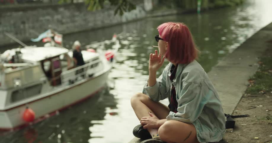 A young woman sits beside a town river smoking a cigarette and waves to a boat passing by