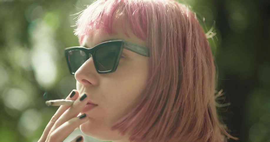 Woman with pink hair and sunglasses smokes a cigarette in public park
