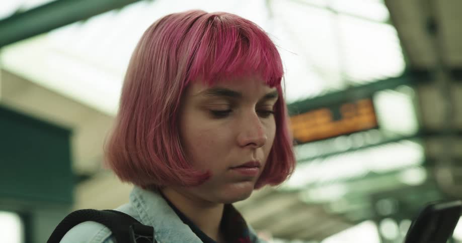 A teenage girl with pink hair is standing in a train station looking around and using her phone