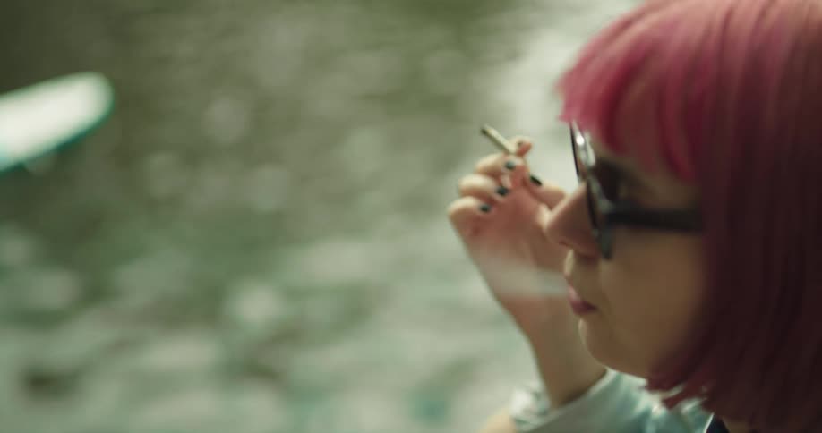 Woman with pink hair and black sunglasses smokes a cigarette and watches body of water