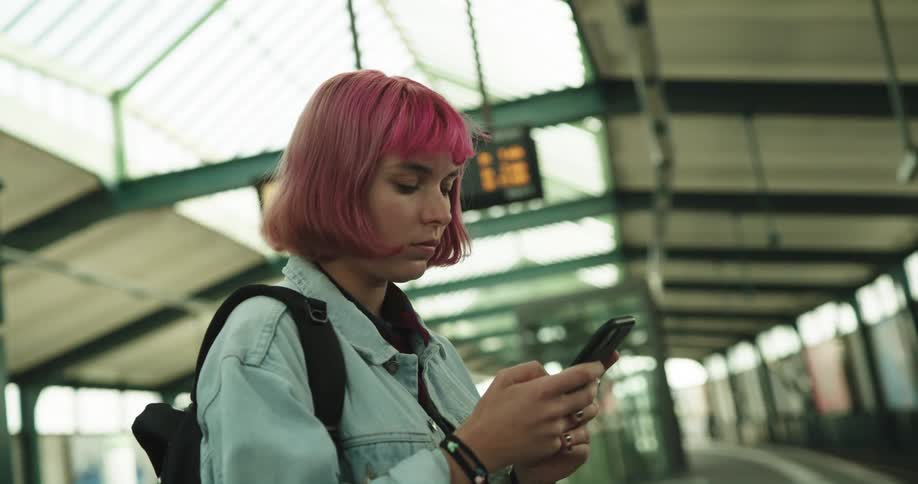 A teenage girl with pink hair is standing in a train station texting on her phone