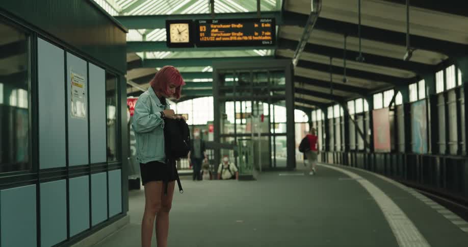 A teenage girl is standing at a train station watching for her train and she takes a book out of her backpack
