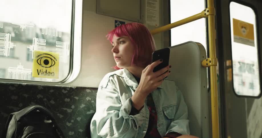 A young woman using her smartphone while riding in a bus stops for a moment to look around at her surroundings before returning her gaze to her phone