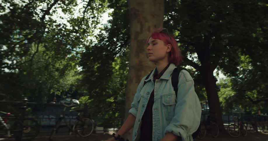A woman with pink hair is riding an electric scooter on a sidewalk in a city park