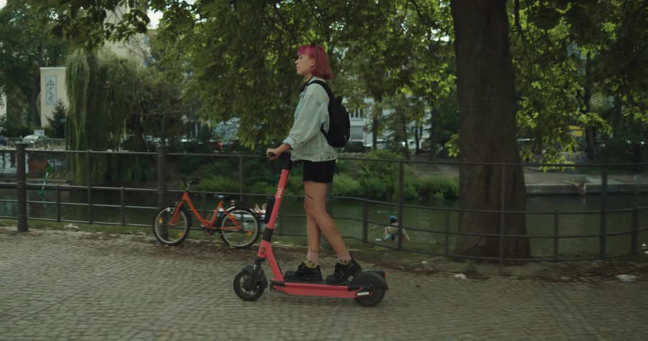 A young woman looks around at her surroundings while riding a motorized scooter along a path next to a waterway during the day