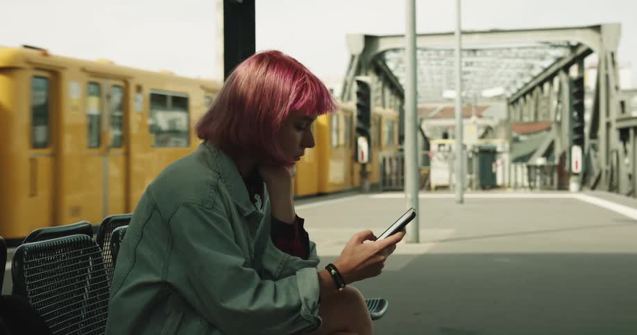 A woman looks at her phone while sitting on a train platform and a train passes