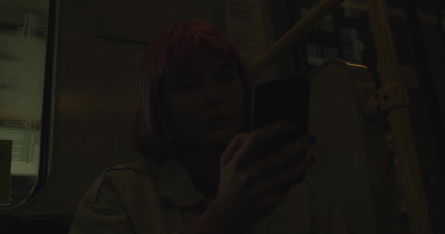 A teenage girl with pink hair is sitting on a train using her cell phone