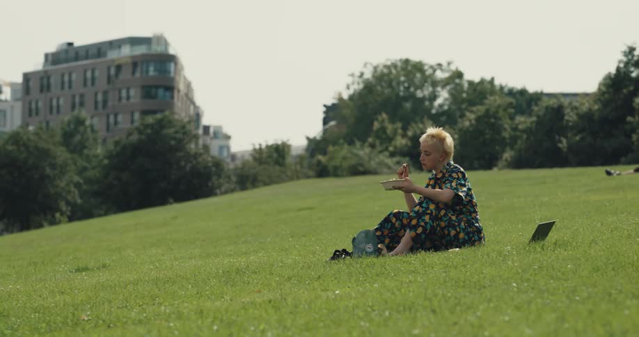 A woman sitting on the grass with her shoes off laptop open eating her lunch in the park
