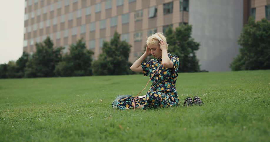 A young woman sitting in a field plugs a pair of headphones into her smartphone before putting the headphones on her head and holding up the phone