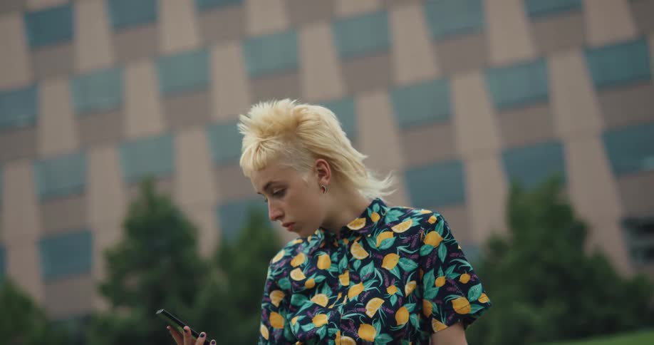 A young woman sitting in a field outside a building checks her smartphone and smiles