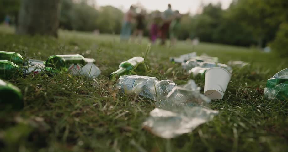 A group of people together sitting in a park throw their empty bottles into the grass before rising to their feet and walking away