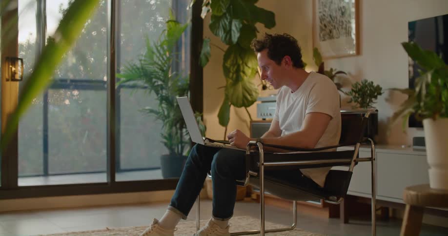 A man is sitting in a chair with a laptop computer having a video conversation with someone and he is talking and laughing
