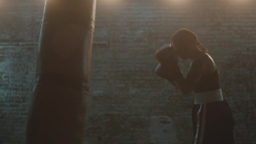 Female boxer with braids sports bra fight trunks and boxing gloves hits puching bag in the gym