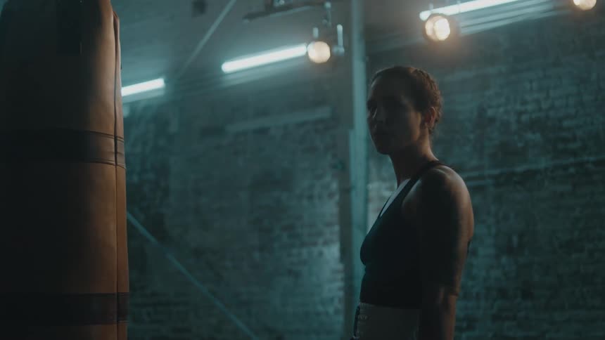 Female boxer with braids sports bra and tattoo stands next to punching bag in the gym