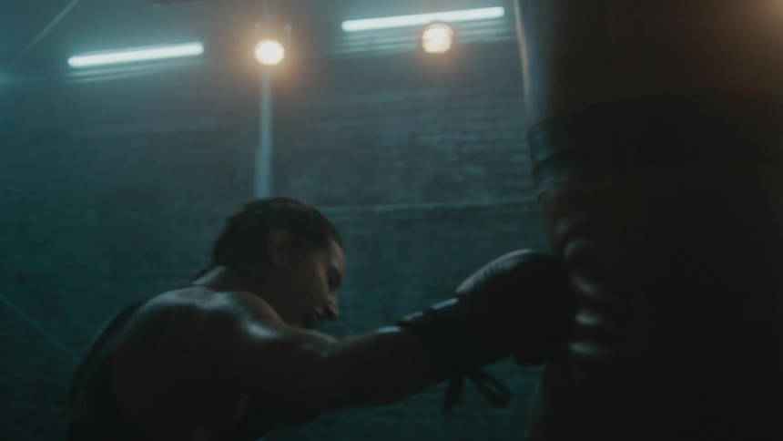 Female boxer with braids wearing a sports bra, fight trunks, and boxing gloves hits punching bag in the gym
