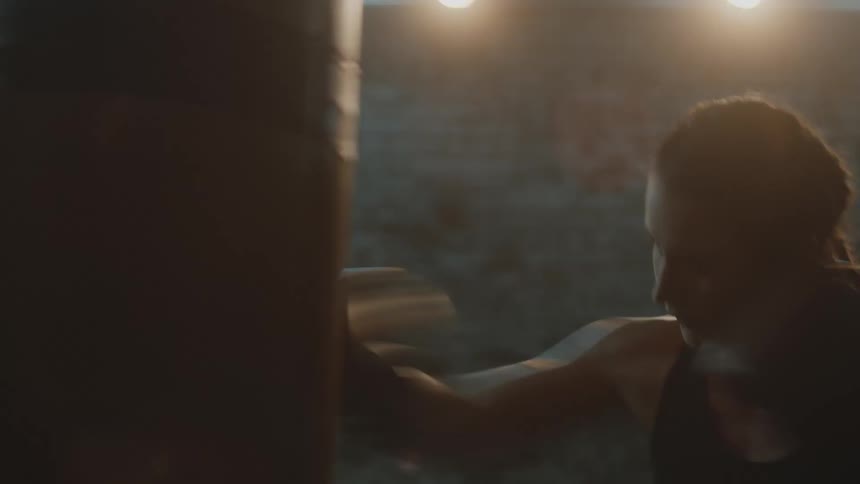 Female boxer with braids sports bra fight trunks and boxing gloves hits puching bag in the gym