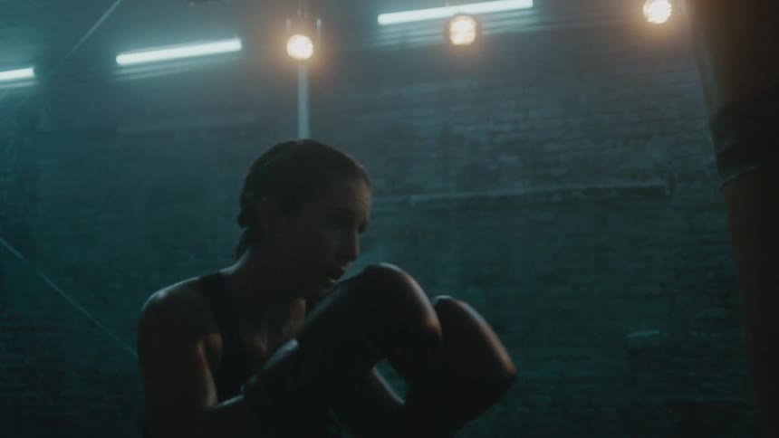 Female boxer with braids wearing a sports bra, fight trunks, and boxing gloves hits punching bag in the gym