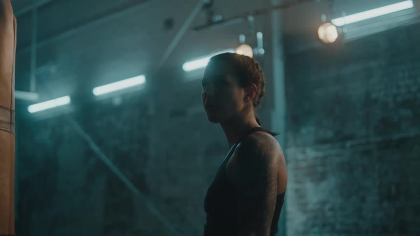 Female boxer with braids sports bra and tattoo stands next to punching bag in the gym