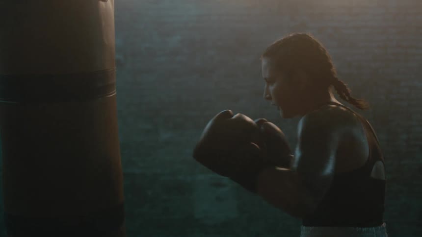 Female boxer with braids wearing a sports bra, fight trunks, and boxing gloves hits punching bag in the gym