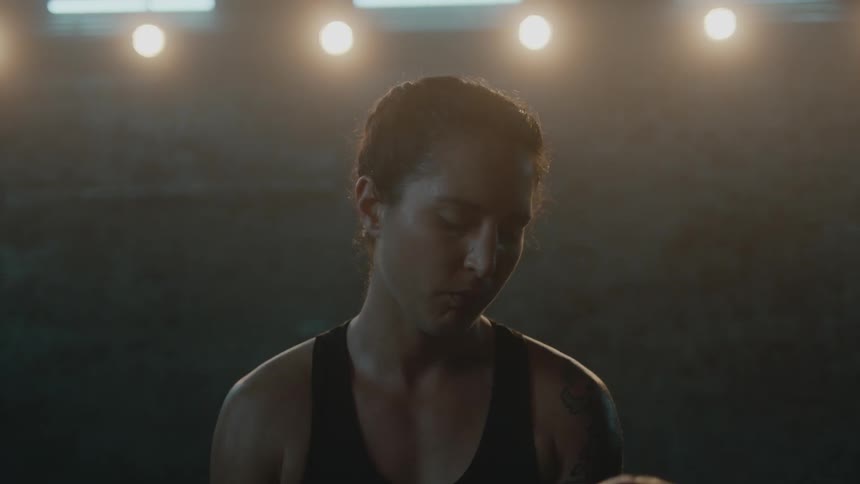 Female athlete takes off boxing gloves and walks away from camera in dimly lit gym