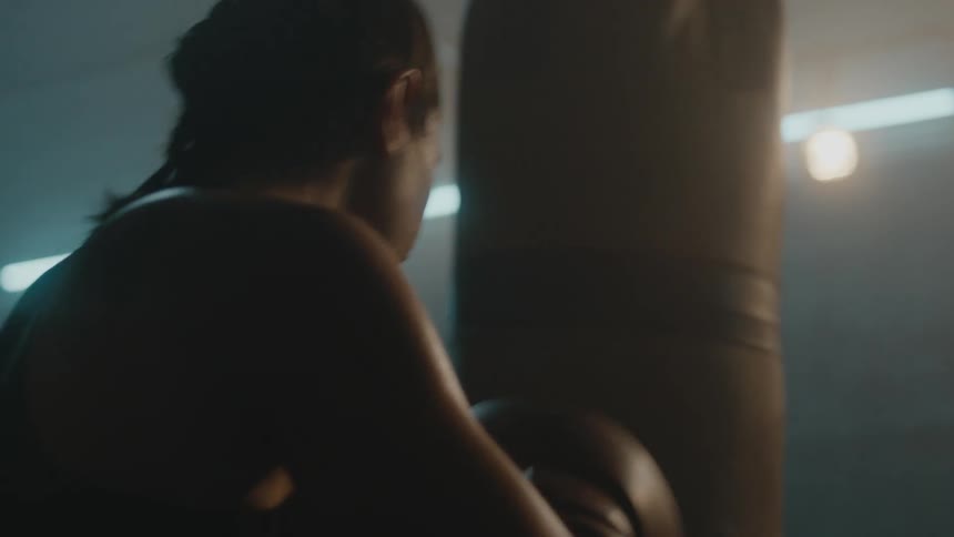 Female boxer with braids sports bra fight trunks and boxing gloves hits puching bag in the gym