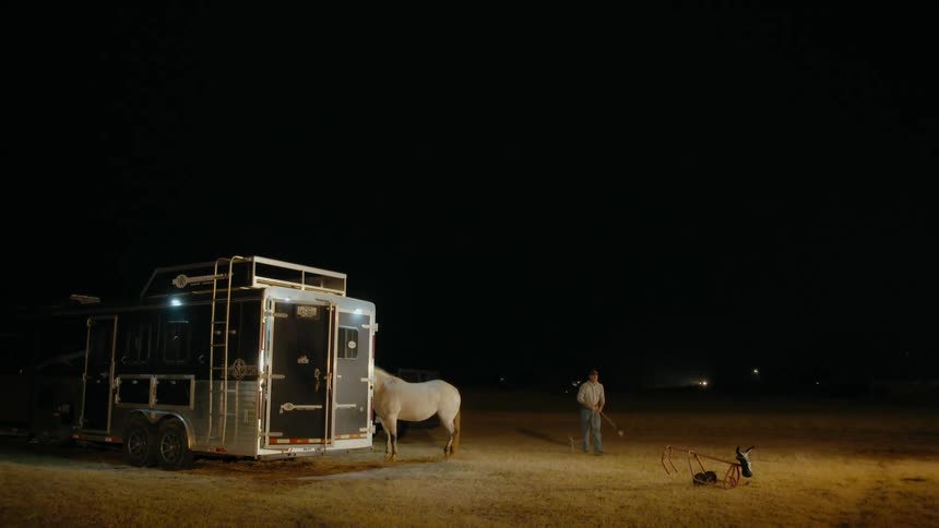 Cowboy practices throwing the lasso near a white horse standing near its transport at night