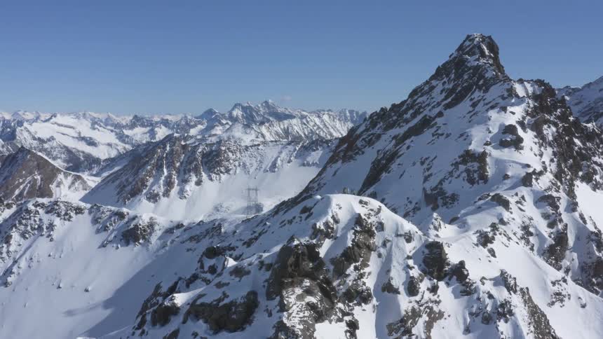The sun shines brightly on a snow covered mountains and there are three people on top one of the peaks