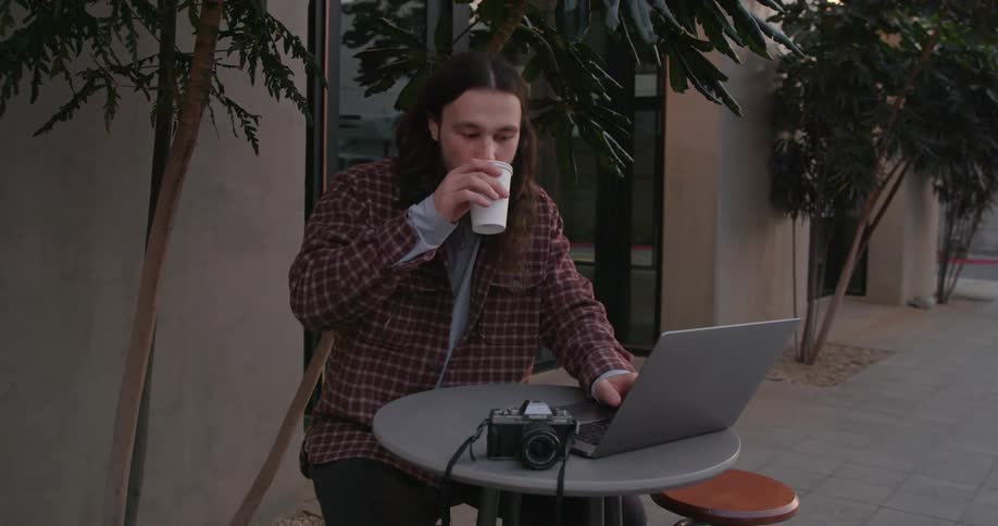 Young long haired man sitting at a table on the city street drinks a beverage while working on a laptop
