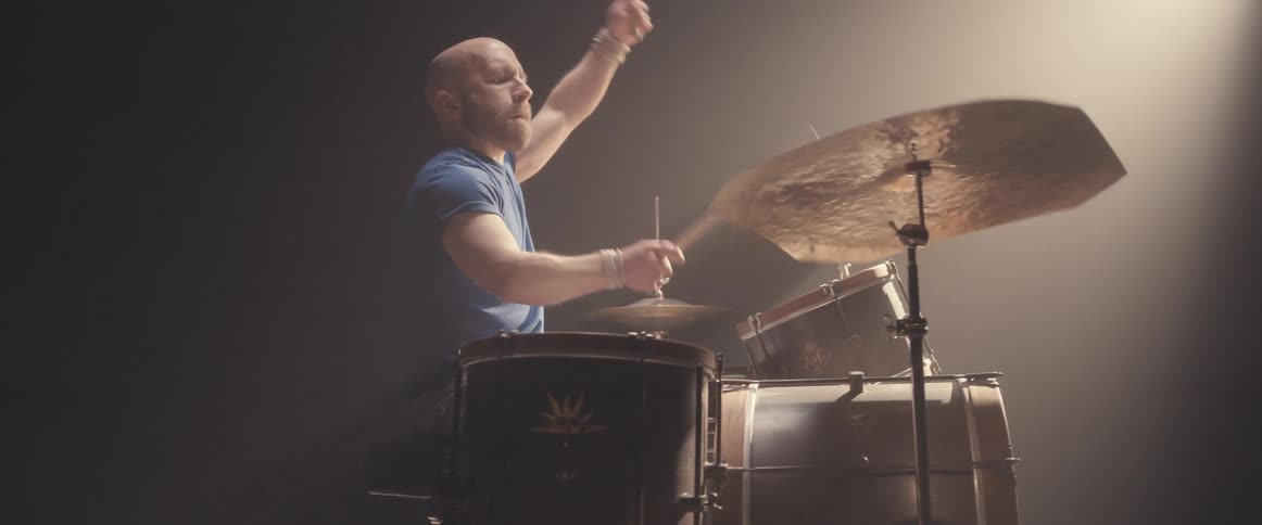 Bald male drummer with blue shirt jeans and beard plays drums in spotlight