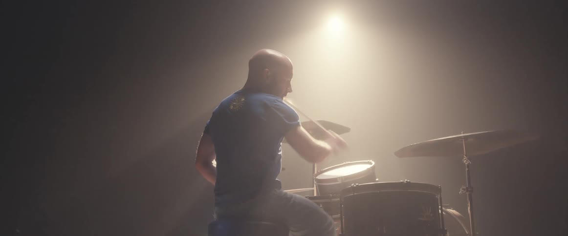 Bald muscular man playing drums on a stage under a spotlight