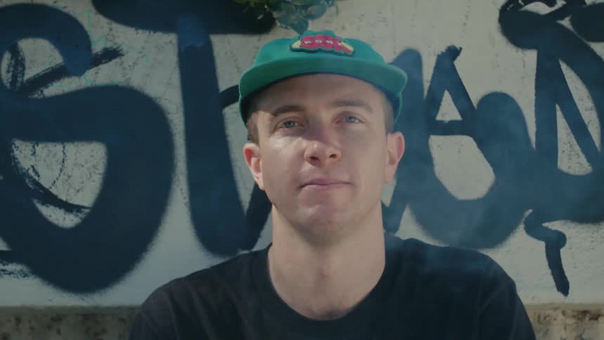 A man smokes a cigarette while posing in front of a graffitied wall