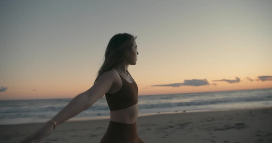 Young woman wearing yoga clothes stretching on the sand beach at sunset next to the ocean