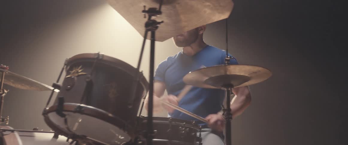 Bald muscular man energetically playing drums on a stage under a spotlight