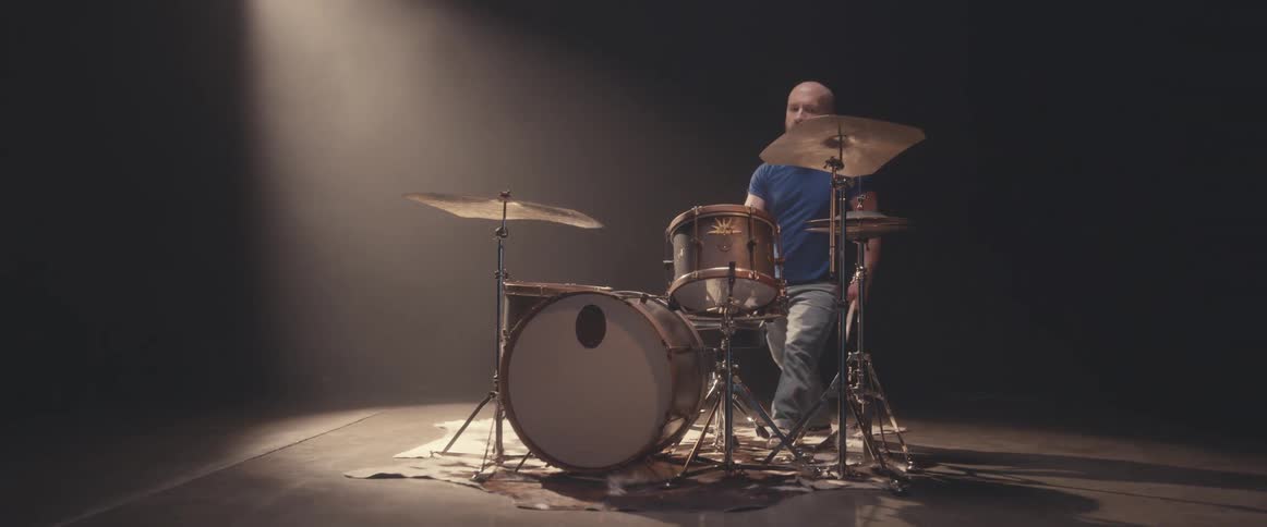 Bald male drummer with blue shirt and jeans and beard plays drums in spotlight