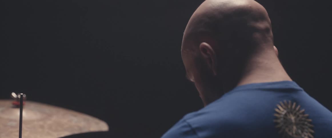 Focused bald male drummer with blue shirt sits behind drum kit in spotlight
