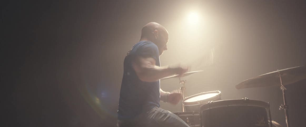Bald male drummer with blue shirt and beard plays drums in spotlight