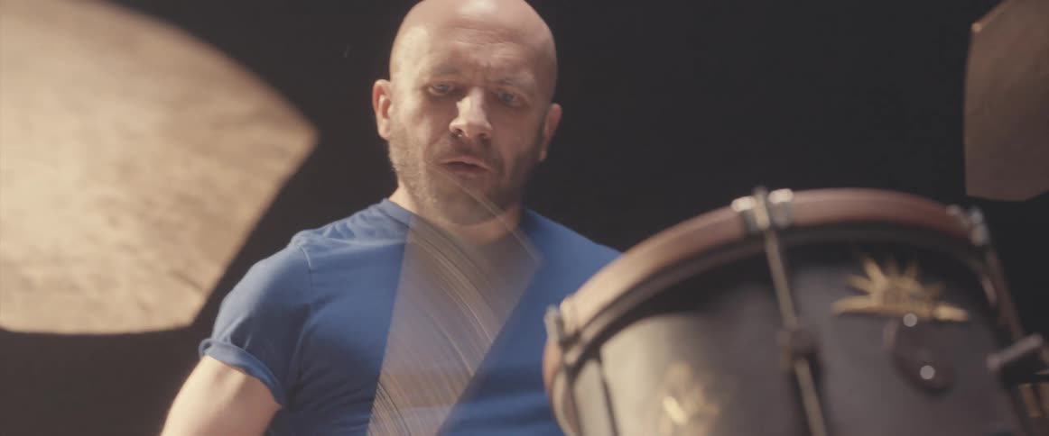 Bald male drummer with blue shirt and beard plays drums in spotlight