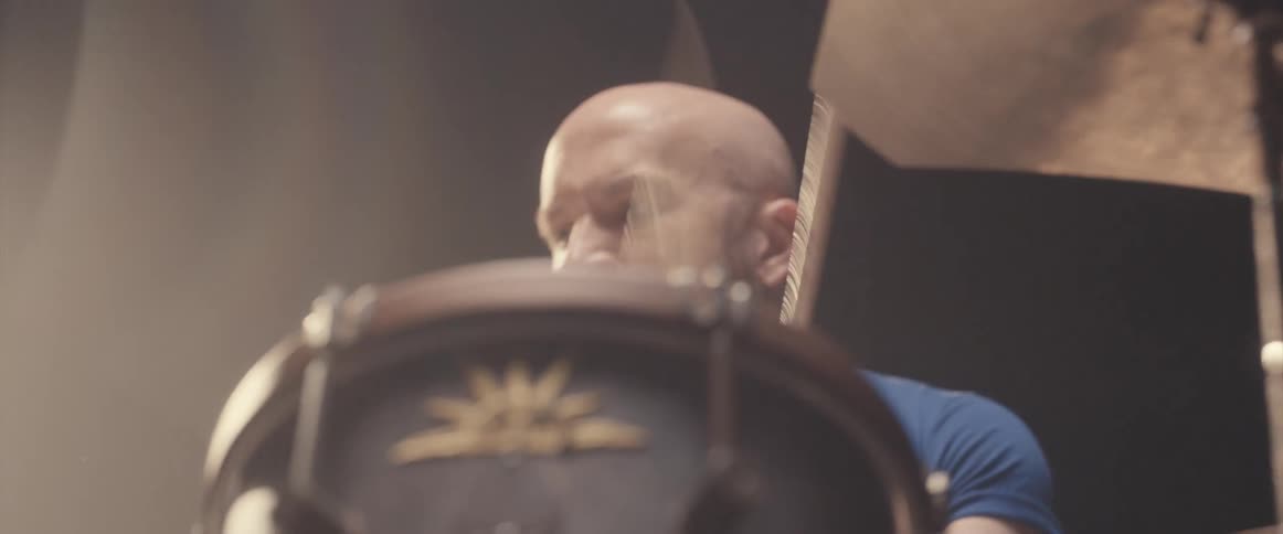 Bald male drummer with blue shirt and beard plays drums in spotlight