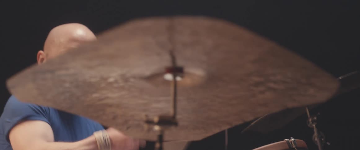 Bald male drummer with blue shirt and beard plays drums in spotlight