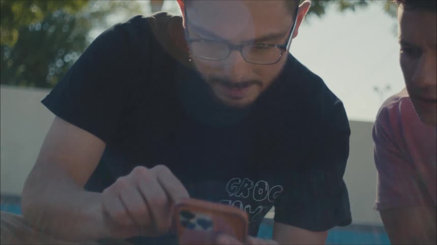 A pair of men sitting outdoors looking at the screen of a smartphone together look to give each other a high five and fist bump