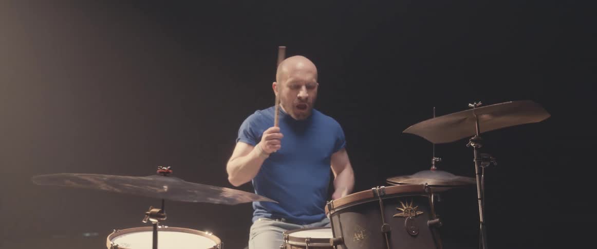 Bald male drummer with blue shirt and jeans and beard plays drums in spotlight