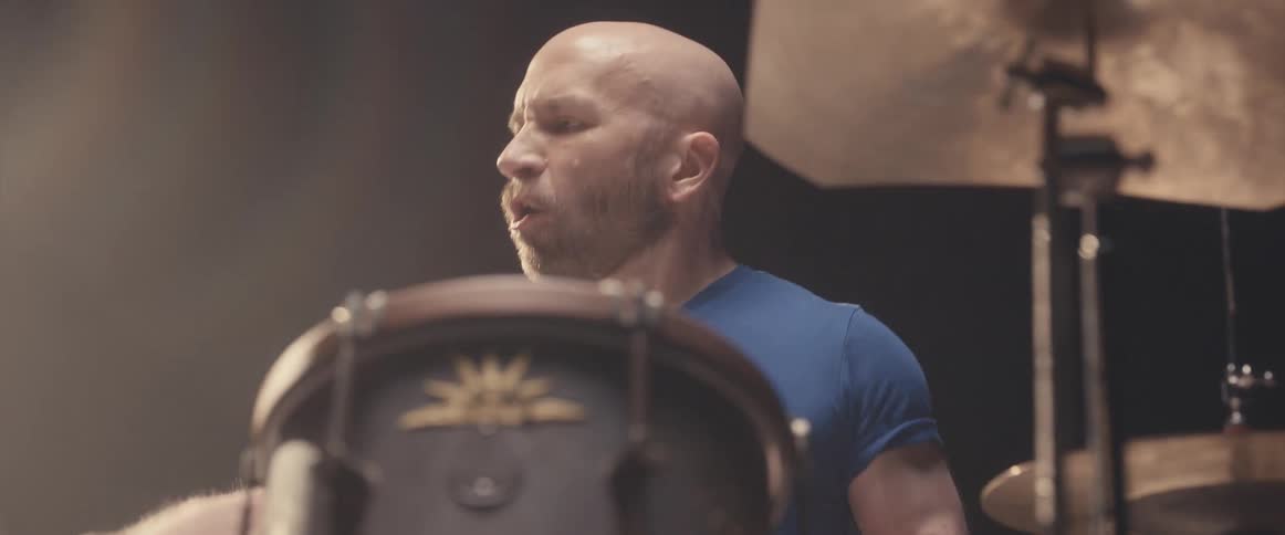 Bald male drummer with blue shirt and beard plays drums in spotlight