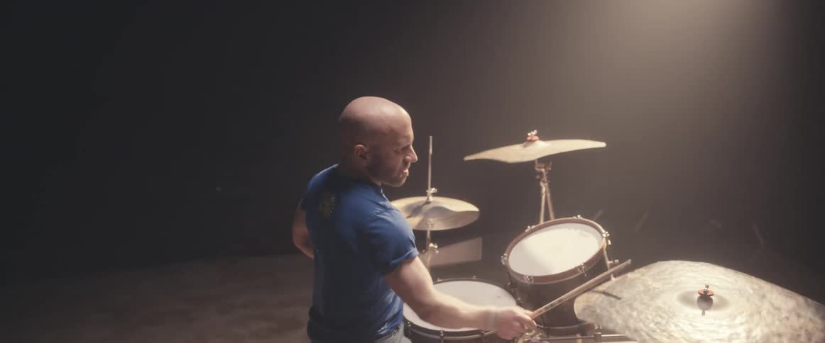 Bald male drummer with blue shirt and beard plays drums in spotlight