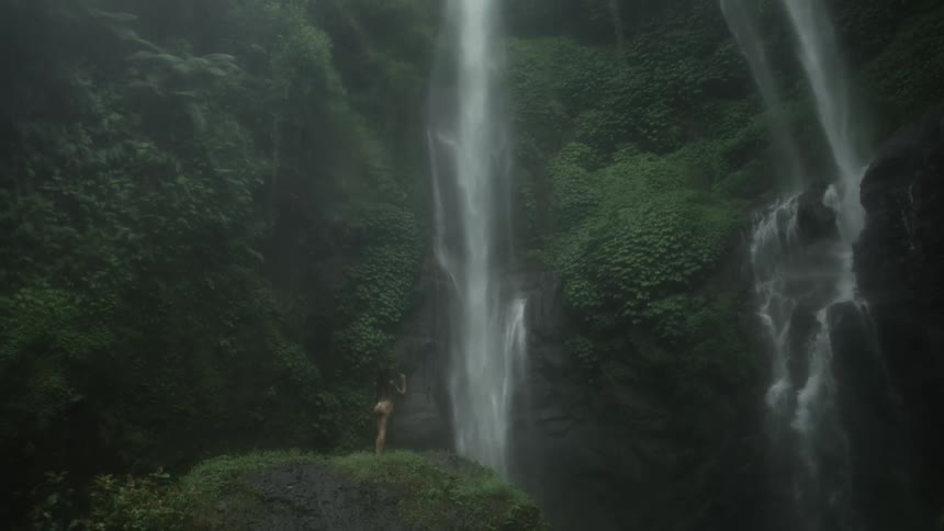 Young fit naked woman stands over a rock covered in green grass in front of waterfalls in the mountain