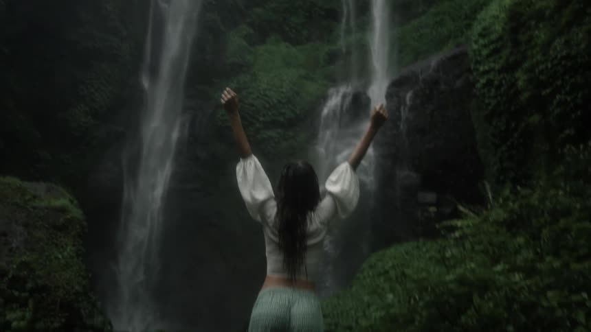 Woman with white blouse green pants raises arms looking at waterfall cascading down cliffside